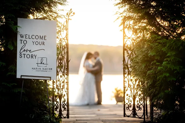 A wedding couple embracing in the background with a 'Welcome to Your Love Story' sign featuring the Hariel Xavier Photography logo in the foreground.