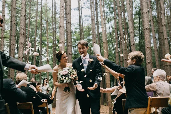 A bride and groom walk down a forest aisle as guests throw flower petals in the air.