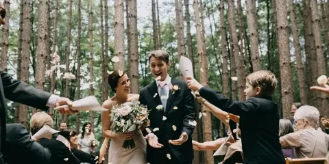 A bride and groom walk down a forest aisle as guests throw flower petals in the air.