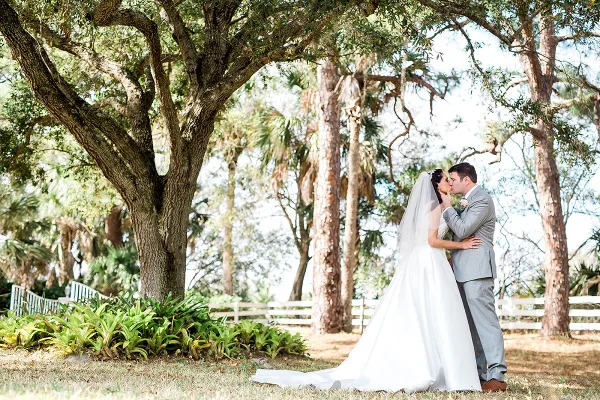 A bride and groom kissing under a large oak tree with a white fence in the background.
