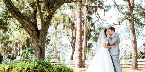 A bride and groom kissing under a large oak tree with a white fence in the background.