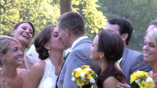 A bride and groom kissing during a wedding ceremony with guests and yellow flowers in the foreground.