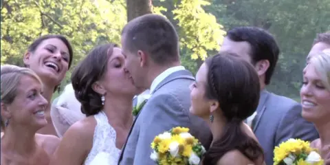 A bride and groom kissing during a wedding ceremony with guests and yellow flowers in the foreground.