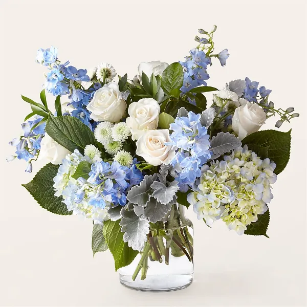 A floral arrangement featuring white roses, blue hydrangeas, and greenery in a clear glass vase.
