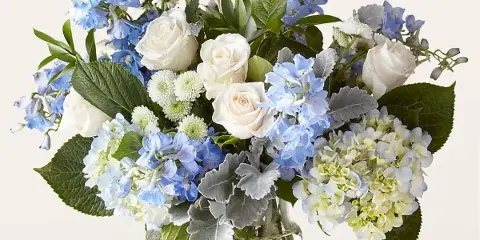 A floral arrangement featuring white roses, blue hydrangeas, and greenery in a clear glass vase.