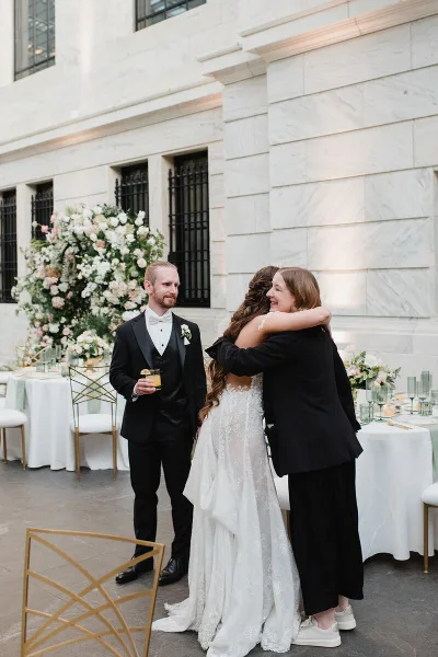 A wedding planner in a black blazer hugs a bride in a white lace gown at a reception, with the groom standing nearby.