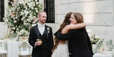 A wedding planner in a black blazer hugs a bride in a white lace gown at a reception, with the groom standing nearby.