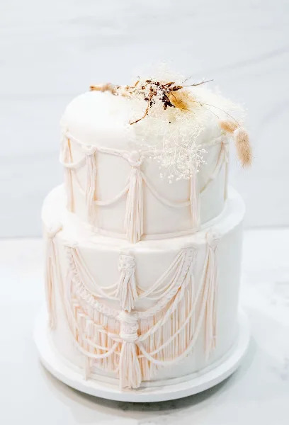 A two-tier white wedding cake decorated with elaborate fondant tassels and dried floral accents.
