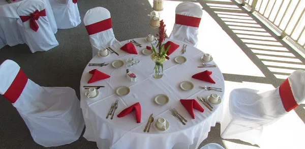 A round banquet table set with white tablecloth, red napkins, silverware, and a floral centerpiece, surrounded by chairs with white covers and red sashes.