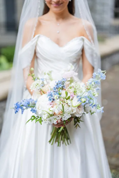 A bride in a white gown holding a lush, garden-style wedding bouquet with white peonies and blue delphiniums.