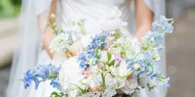 A bride in a white gown holding a lush, garden-style wedding bouquet with white peonies and blue delphiniums.
