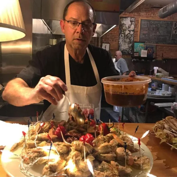 Chef Peter Hadley in a white apron plating a large dish of appetizers in a commercial kitchen.