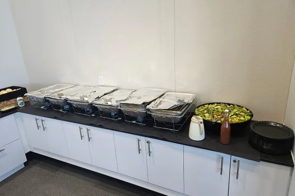 A catering buffet line featuring foil-covered chafing dishes, a large salad bowl, and stacked plates set up on a counter.