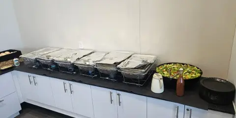 A catering buffet line featuring foil-covered chafing dishes, a large salad bowl, and stacked plates set up on a counter.