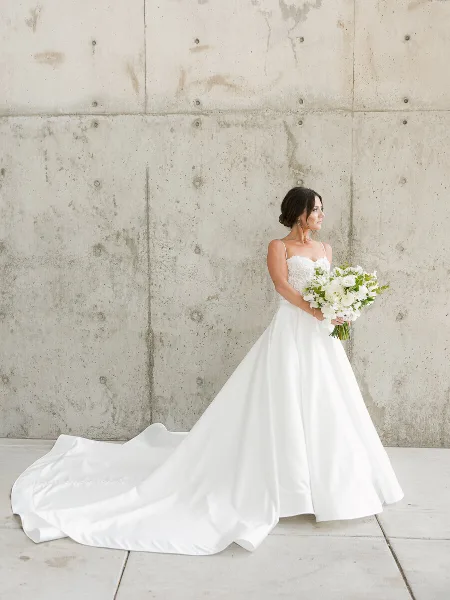 A bride in a white wedding dress holding a bouquet of white flowers standing against a concrete wall.