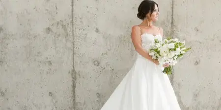 A bride in a white wedding dress holding a bouquet of white flowers standing against a concrete wall.