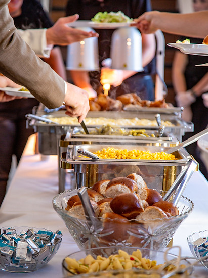 A catering buffet line featuring chafing dishes with food, a bowl of bread rolls, and appetizers at an event.
