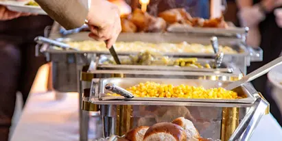 A catering buffet line featuring chafing dishes with food, a bowl of bread rolls, and appetizers at an event.