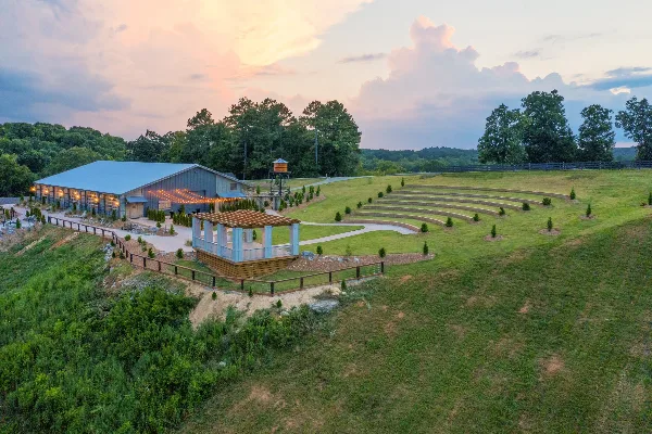 Aerial view of a wedding venue featuring a large barn-style building, an outdoor ceremony pergola, and tiered grass seating on a hillside.