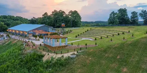 Aerial view of a wedding venue featuring a large barn-style building, an outdoor ceremony pergola, and tiered grass seating on a hillside.