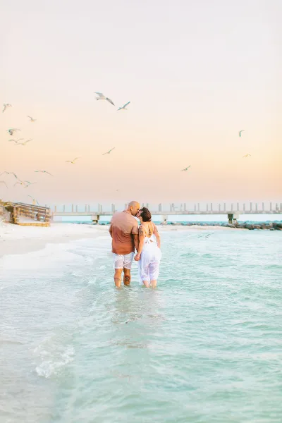 A couple standing in shallow ocean water on a beach at sunset with seagulls flying overhead.