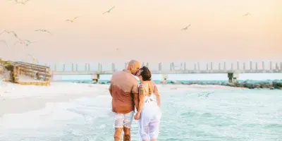 A couple standing in shallow ocean water on a beach at sunset with seagulls flying overhead.