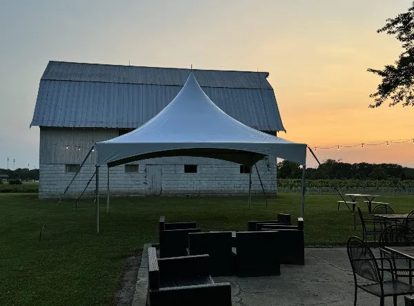 A white event tent set up on a grassy field at dusk with outdoor seating and string lights, in front of a barn-style building.