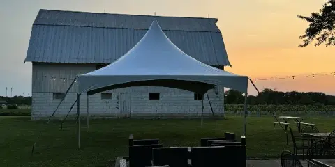 A white event tent set up on a grassy field at dusk with outdoor seating and string lights, in front of a barn-style building.