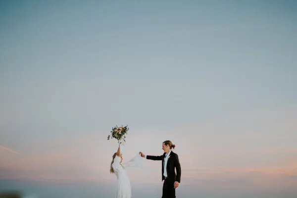 A bride and groom standing on a beach at dusk, with the bride holding a bouquet and the groom holding her hand.