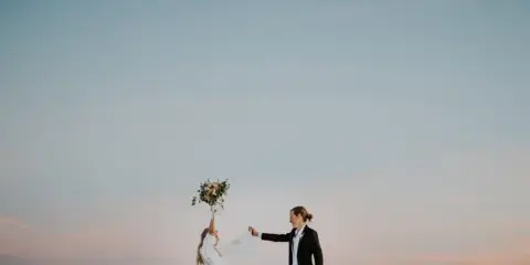 A bride and groom standing on a beach at dusk, with the bride holding a bouquet and the groom holding her hand.