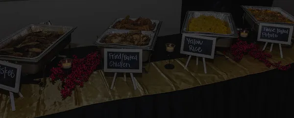 A catering buffet table featuring chafing dishes with fried chicken, yellow rice, and twice-baked potatoes, decorated with red berries and small chalkboard signs.