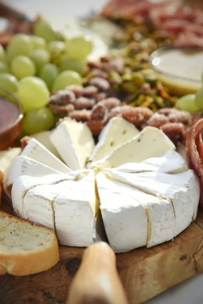 A close-up of a catering spread featuring a wheel of sliced brie cheese, green grapes, and nuts on a wooden board.