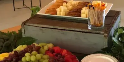 A catering buffet table featuring a platter of cheese cubes and crackers, alongside a bowl of fresh fruit including grapes, watermelon, and pineapple.