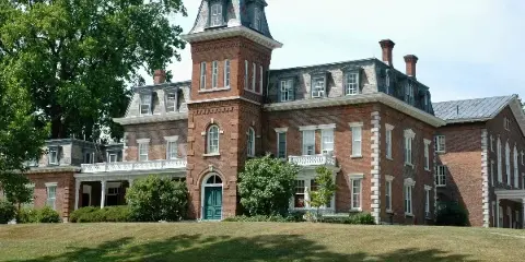 Exterior view of the Oneida Community Mansion House, a historic red brick building with a central tower and white balconies, set on a grassy lawn.