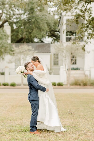 A bride and groom embracing outdoors in a grassy area with a white building in the background.