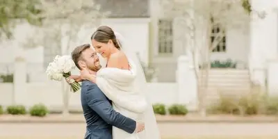 A bride and groom embracing outdoors in a grassy area with a white building in the background.