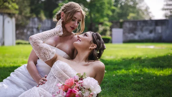 Two women in white wedding dresses posing affectionately on a grassy lawn with a bouquet of flowers.