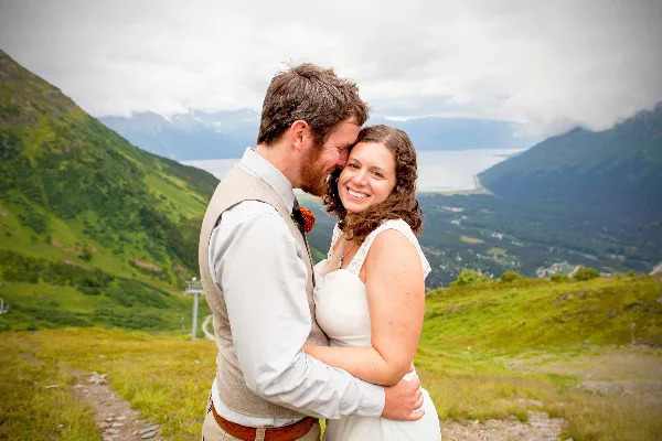 A bride and groom embracing in a white dress and vest against a backdrop of green mountains and a lake.