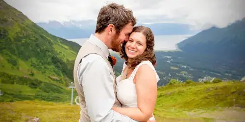 A bride and groom embracing in a white dress and vest against a backdrop of green mountains and a lake.