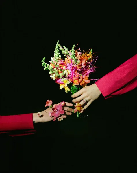 A colorful, whimsical floral bouquet held by two hands against a black background, featuring pink, orange, and green blooms.