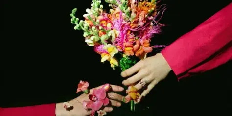 A colorful, whimsical floral bouquet held by two hands against a black background, featuring pink, orange, and green blooms.