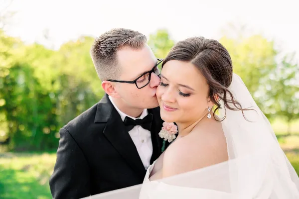 A groom in a black tuxedo kissing the forehead of a bride wearing a white veil and dress, set against a blurred green outdoor background.