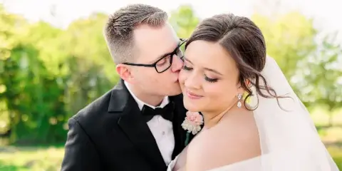A groom in a black tuxedo kissing the forehead of a bride wearing a white veil and dress, set against a blurred green outdoor background.