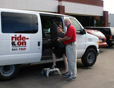 A driver in a red shirt assists a passenger boarding a white van branded with the Ride-On Transportation logo.