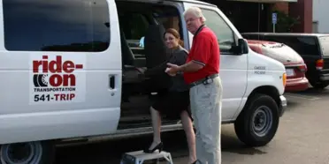 A driver in a red shirt assists a passenger boarding a white van branded with the Ride-On Transportation logo.