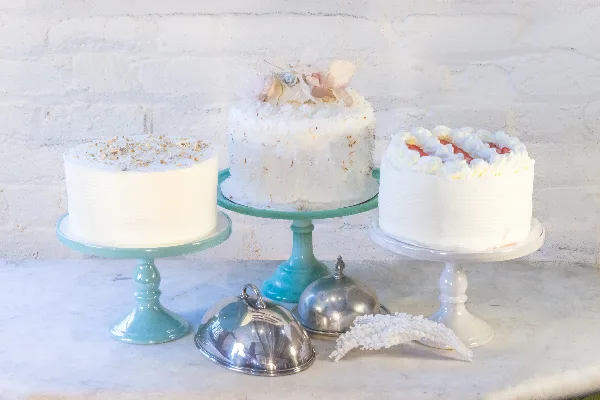 Three decorated cakes displayed on pastel-colored cake stands against a white brick wall.
