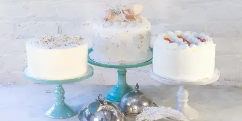 Three decorated cakes displayed on pastel-colored cake stands against a white brick wall.