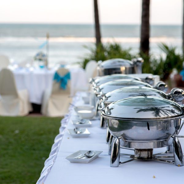 A row of silver chafing dishes set up on a white tablecloth at an outdoor beachside event.