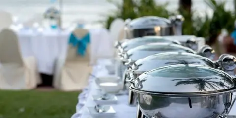 A row of silver chafing dishes set up on a white tablecloth at an outdoor beachside event.