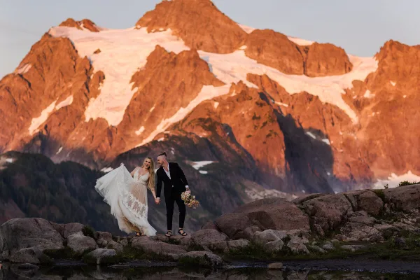 A bride and groom standing on a rocky mountain peak with a large snow-capped mountain in the background.